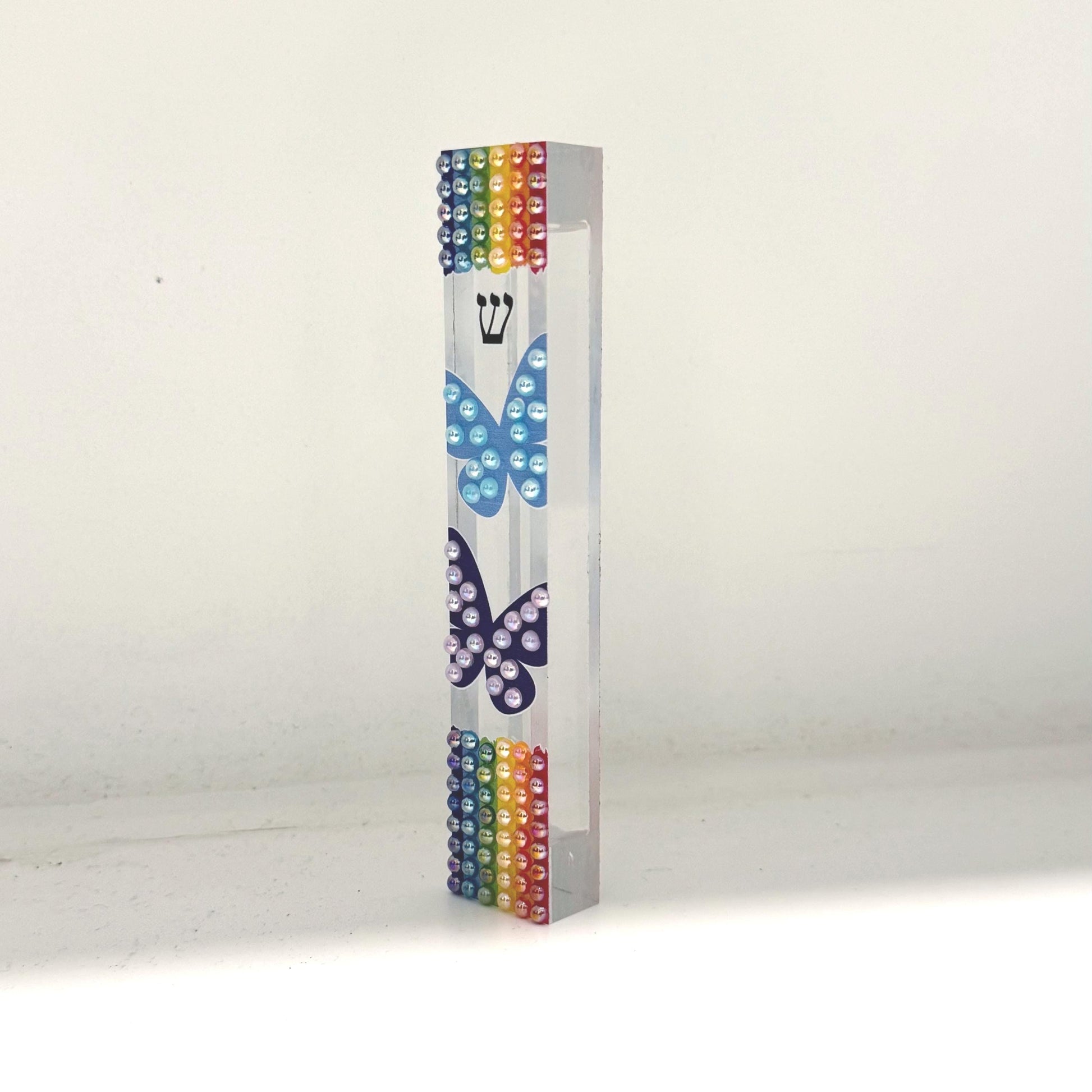 May include: A clear acrylic mezuzah case with rainbow-colored beads at the top and bottom. Two butterfly designs are painted on the front, one light blue and one dark blue. A Hebrew letter shin is visible above the light blue butterfly.